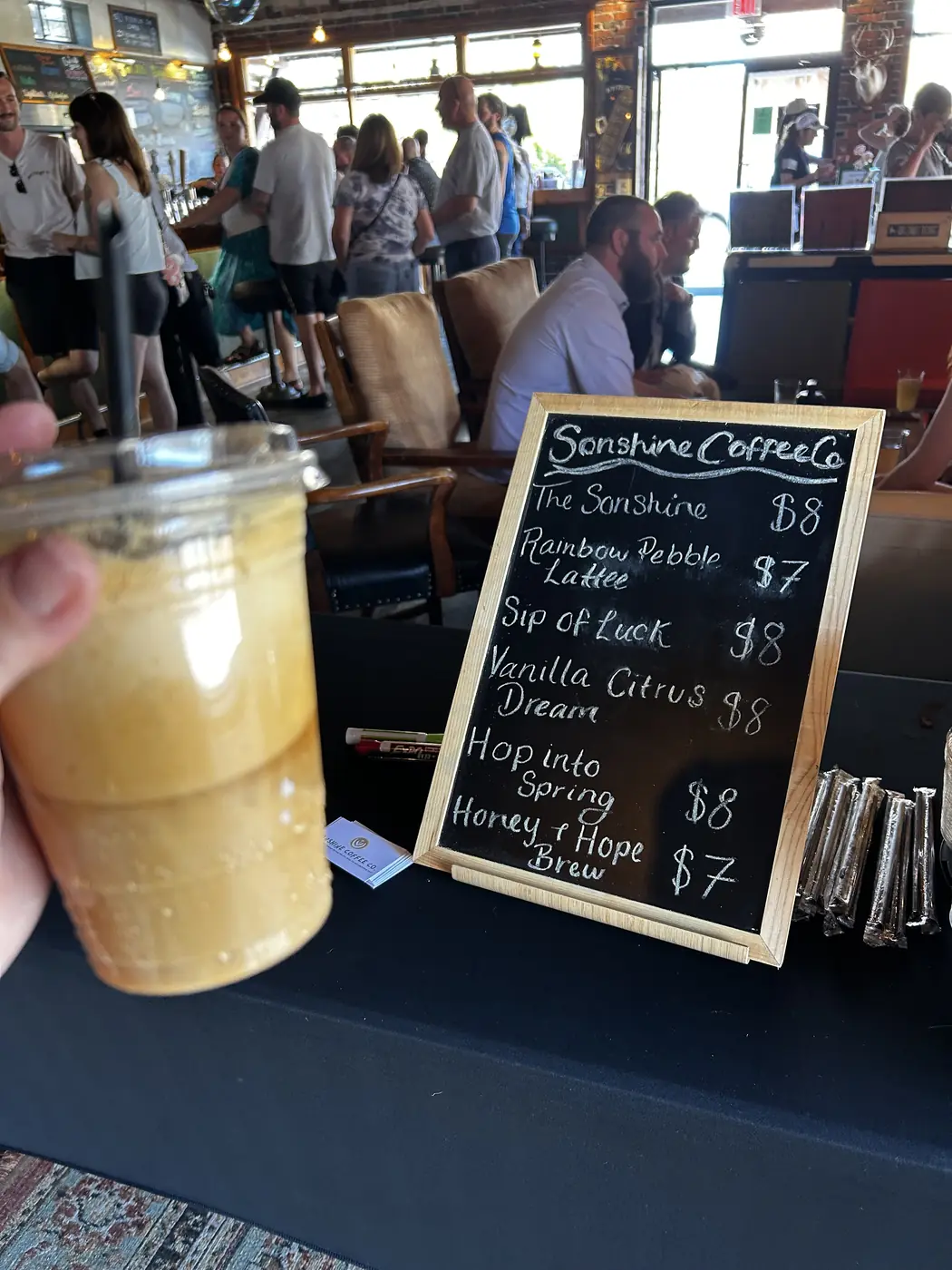 An iced coffee held up in front of the chalkboard menu at a busy indoor event.