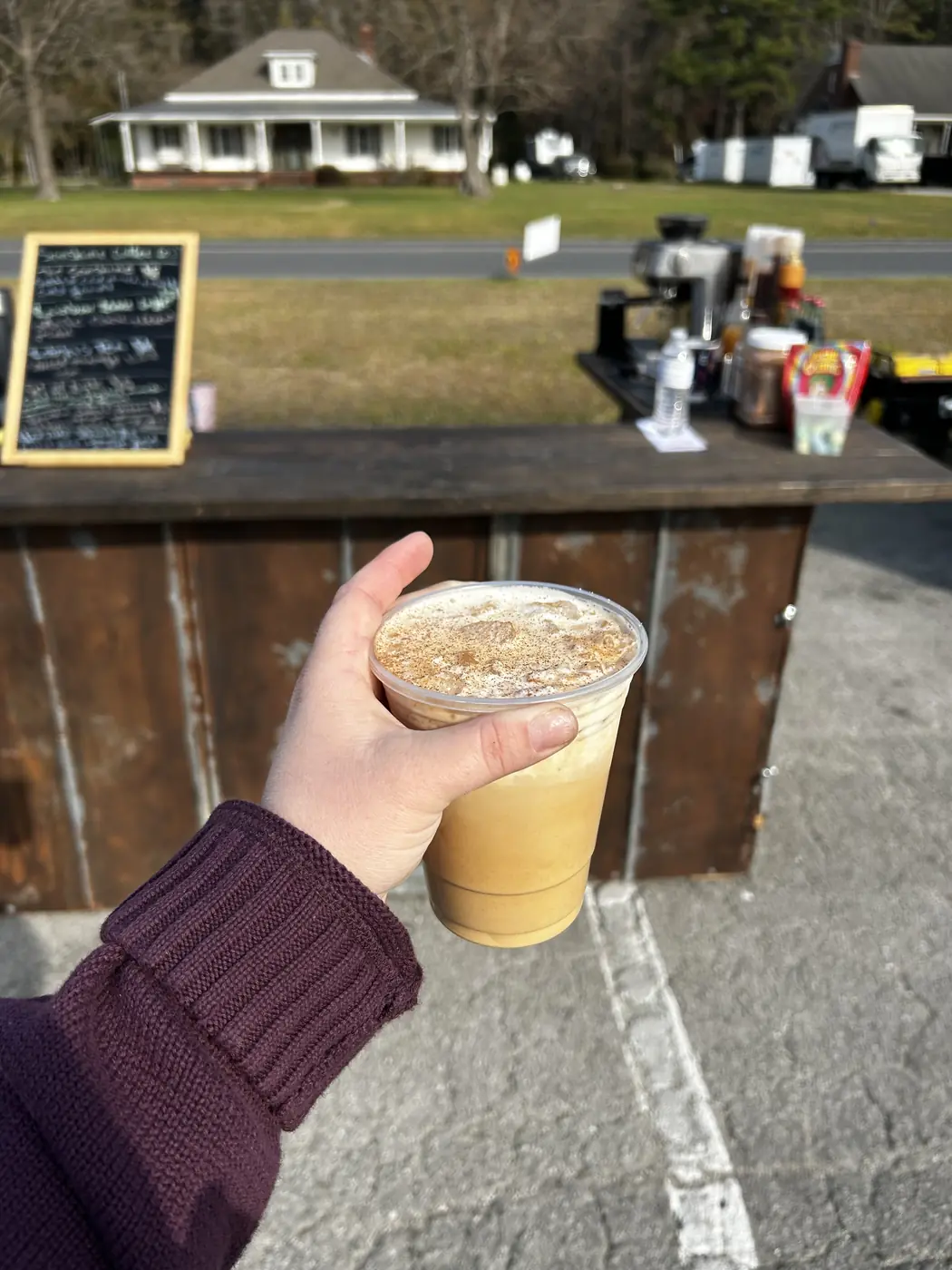 An iced coffee held in front of the wooden cart at an outdoor event.
