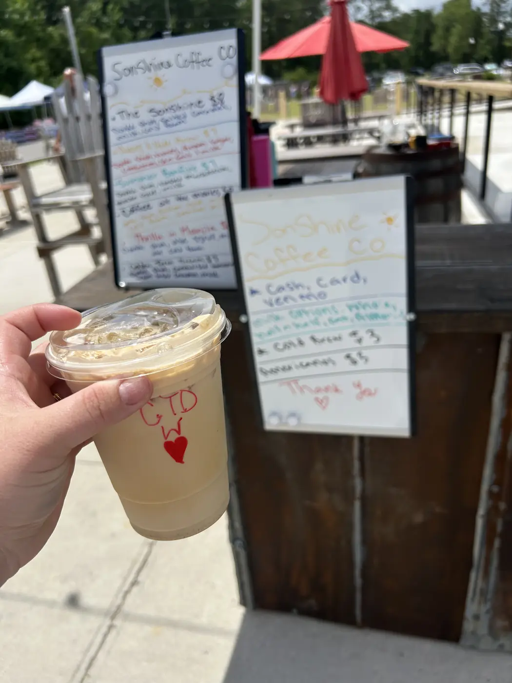 An iced latte with a heart drawn on the lid, handwritten menus behind it at an outdoor market.
